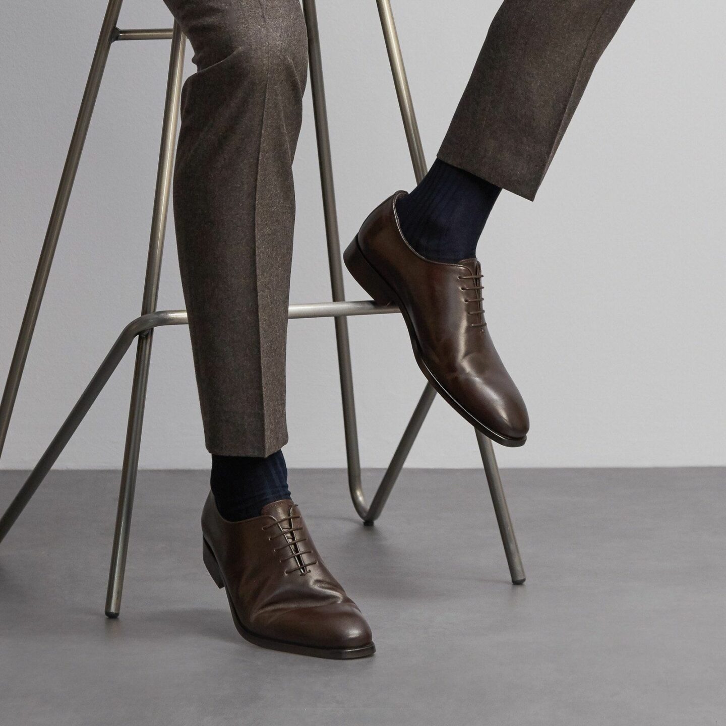 Close up of a man's legs sitting in a grey suit, navy sartorial socks and brown shoes