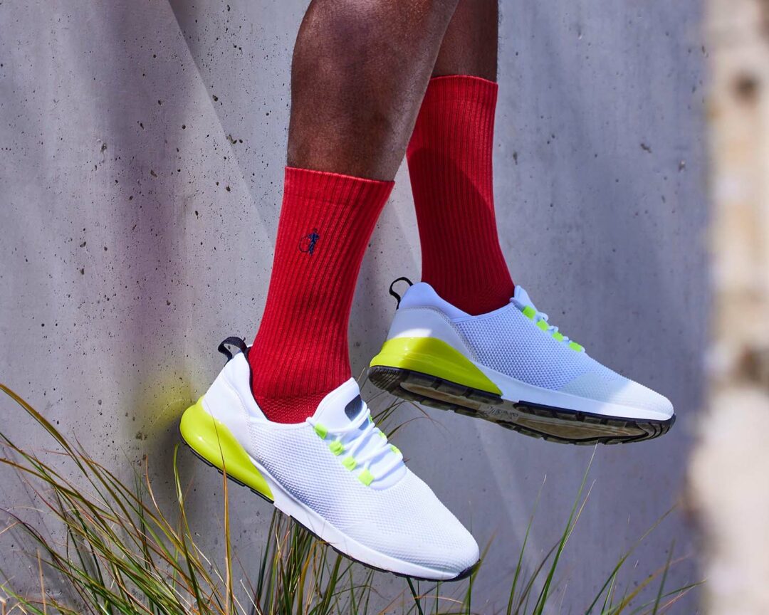 A man in white sneakers and red socks from the London Sock Company leaning against a wall.