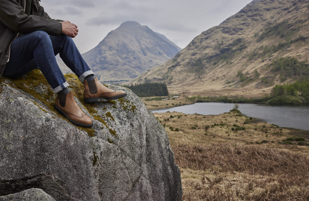 A man sits on a rock with a view of a Highland glen behind him, wearing jeans, Chelsea boots and grey boot socks by London Sock Company.