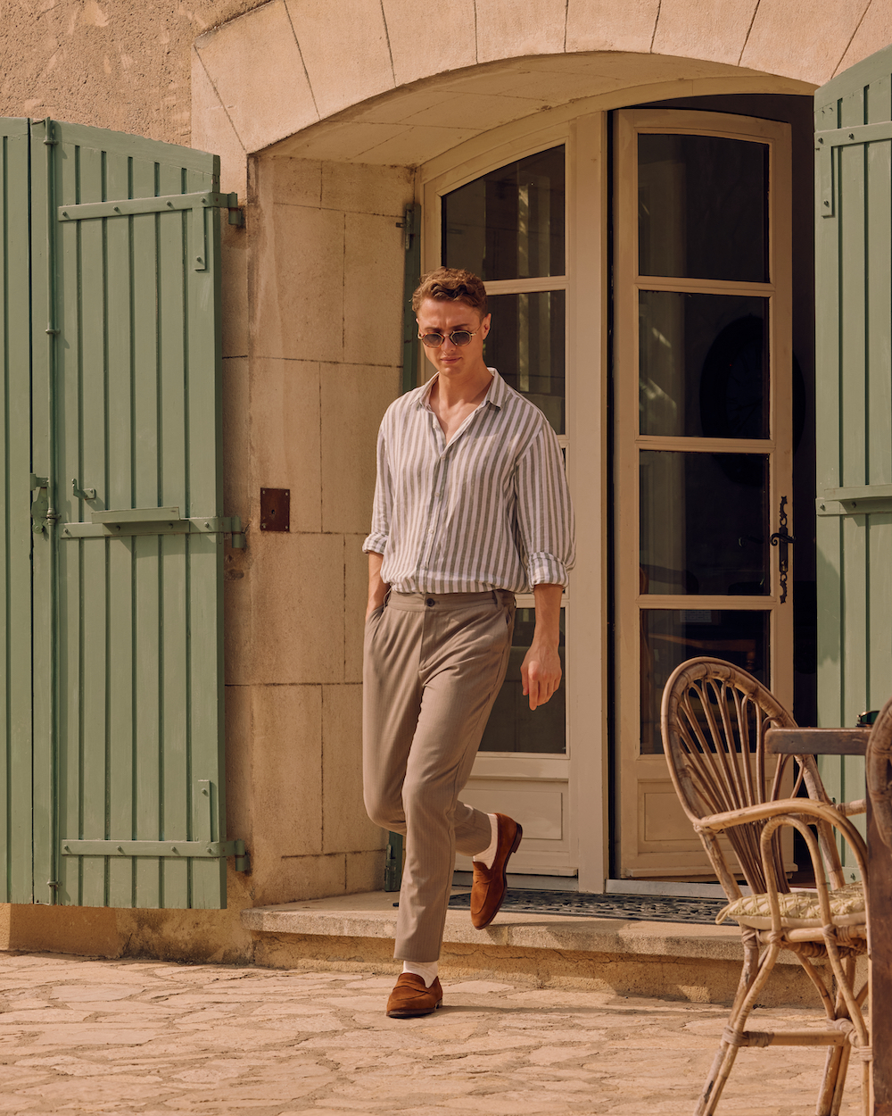 A man wearing tan chinos and a striped linen shirt, walks out of a shuttered doorway. On his feet are brown suede loafers and white socks by London Sock Company.